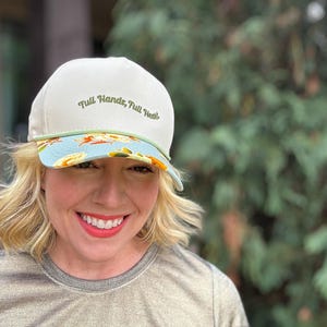 May include: A white baseball cap with the embroidered text "Full Hands, Full Heart." The brim features a floral pattern with blue, yellow, and orange accents. A green rope detail accents the cap.