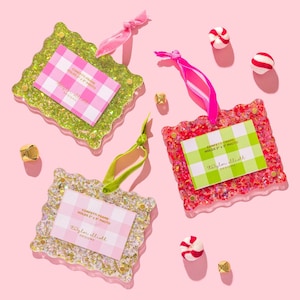 May include: Three decorative confetti frames in green, red, and gold, each designed to hold a 5 cm x 7.6 cm photo. The frames have scalloped edges and are embellished with ribbons. The image also features sweets and small gold bells on a pink backdrop.