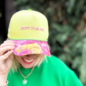 Op de afbeelding: Een gele baseballcap met de roze tekst "HAPPY TO BE HERE". De klep heeft een kleurrijk bloemenpatroon in roze, geel en oranje. De persoon draagt een groene trui.
