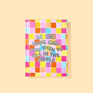 May include: A colorful notebook with a grid pattern of pink, orange, yellow, and blue squares. The cover features the text "BE THE HYPE GIRL YOU WISH TO SEE IN THE WORLD" in glittery silver letters.