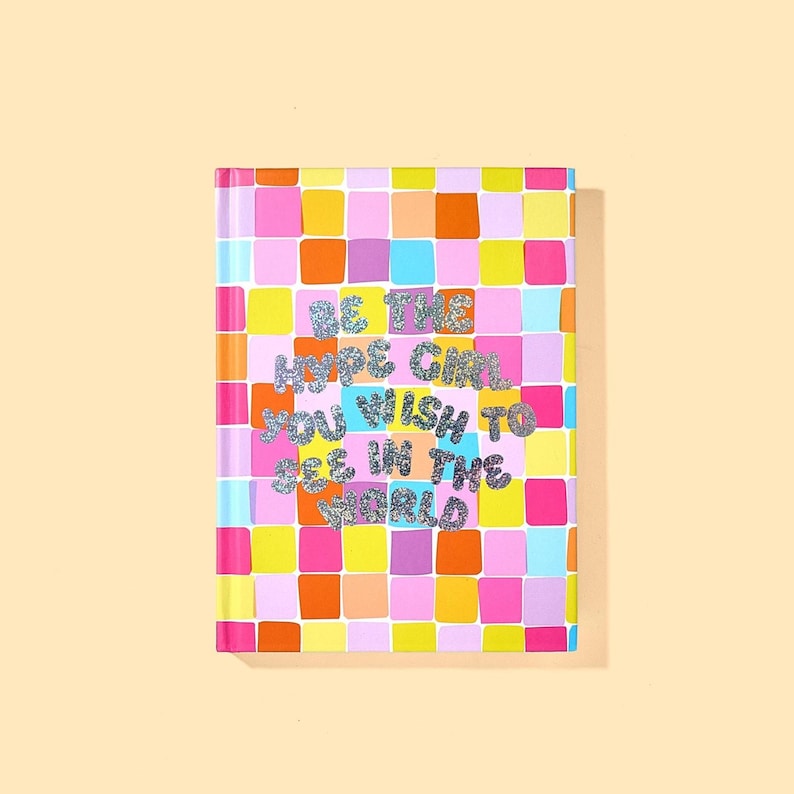 May include: A colorful notebook with a grid pattern of pink, orange, yellow, and blue squares. The cover features the text "BE THE HYPE GIRL YOU WISH TO SEE IN THE WORLD" in glittery silver letters.