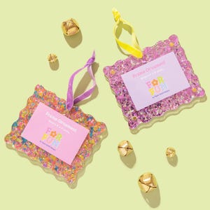 May include: Two decorative frame ornaments with the text "FOR FUN" on a pink card. The frames are pink and multi-coloured with a wavy edge. Each ornament has a ribbon for hanging. Gold bells are scattered around the ornaments.