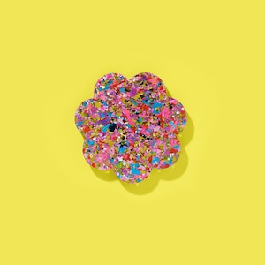 Puede incluir: Un accesorio en forma de flor con un patrón de confeti en tonos rosa, azul y amarillo. El objeto se encuentra sobre un fondo amarillo brillante, creando un contraste vibrante. El diseño del accesorio evoca un estilo lúdico y artístico.