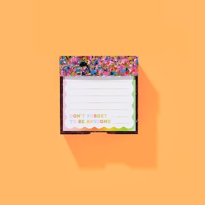 Peut inclure: Un bloc-notes rectangulaire avec une bordure supérieure colorée en forme de confettis et le texte "DON'T FORGET TO BE AWESOME" en couleurs arc-en-ciel. Le bloc-notes a des pages lignées et un bord festonné, sur fond orange.