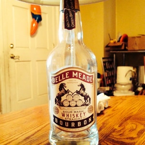 Könnte beinhalten: Eine leere Belle Meade Bourbon-Flasche mit einem Etikett mit zwei sich aufbäumenden Pferden. Das Etikett enthält auch den Text "Sour Mash Whiskey Straight Bourbon". Die Flasche ist klar und steht auf einer Holzoberfläche.