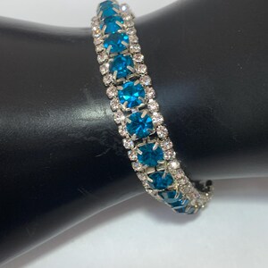 May include: A silver-toned bracelet with alternating rows of clear and teal-colored gemstones. The bracelet is set on a black surface, showcasing the sparkle of the gems. This piece of jewelry is suitable for special occasions.