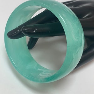 May include: A wide, translucent bangle bracelet in shades of teal and white. The marbled effect gives the bracelet a unique appearance. The bracelet is a solid, circular shape and appears to be made of a smooth material.