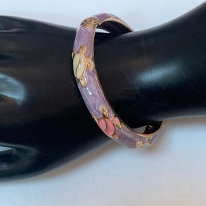 May include: A hinged bangle bracelet featuring a lavender enamel base with gold-tone metal edges. The surface is decorated with small, colorful turtle motifs in shades of cream, pink, and green, arranged in a repeating pattern around the band.