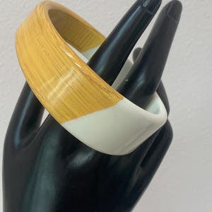 May include: A chunky, two-tone bangle bracelet. The top half is a light yellow with a wood grain pattern, and the bottom half is white. The bracelet is displayed on a black hand.