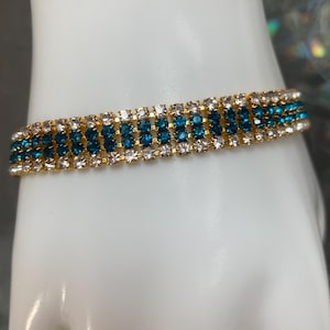 May include: A gold-toned bracelet with three rows of sparkling gemstones. The center row features teal-colored stones, while the top and bottom rows are adorned with clear, white stones. The bracelet is displayed on a white surface.
