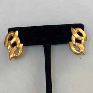 May include: A pair of gold-toned earrings featuring a stylized, interlocking chain-link design. Each earring has a smooth, matte finish with curved, rounded edges that create an open, geometric shape. The earrings are symmetrical in their construction.