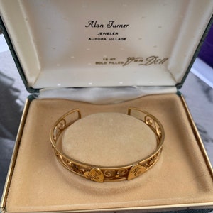 May include: A gold-filled bracelet with heart-shaped accents and the names "Mint" and "Ma" engraved on it, displayed in a vintage jewelry box. The box is green with the jeweler's name, "Alan Turner," printed on the inside.