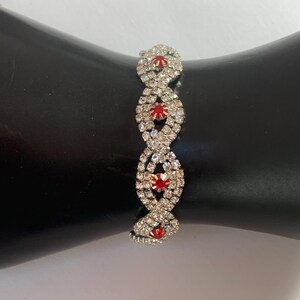 May include: A silver-toned bracelet with a series of oval-shaped links. Each link is encrusted with clear rhinestones, surrounding a central red gemstone. The bracelet is displayed against a black background.