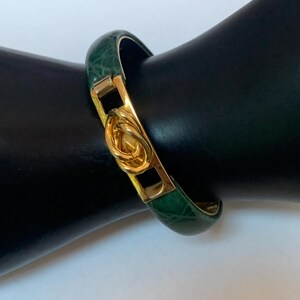 May include: A hinged bangle bracelet featuring a dark green, textured, faux-alligator leather inlay. The bracelet is accented with a polished gold-tone metal center piece that displays an interlocking, looped knot design and a secure clasp mechanism.