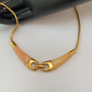 May include: A gold-toned necklace with a coral-colored center and a snake chain. The necklace features a curved design with a gold clasp. The coral-colored center is a light peach color.