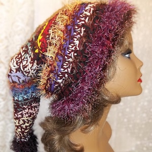 May include: A handmade, colorful, long-tail hat. The hat features a mix of burgundy, purple, yellow, and white yarns, with a fluffy, fringed trim. The hat is displayed on a mannequin head.