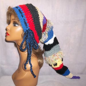 May include: A colorful, handmade crochet hat with a long, pointed tail. The hat features a variety of colors, including blue, red, black, and beige, and is adorned with blue tassels. The hat is designed to be worn on the head.