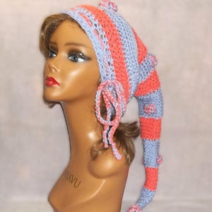 May include: A handmade, whimsical crochet hat in coral and light blue. The hat features vertical stripes, a long, pointed tail, and decorative pom-poms. The hat is displayed on a mannequin head.