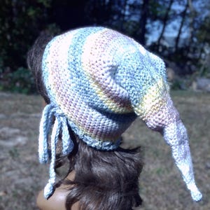 May include: A crochet hat featuring a long, pointed tail and a pastel color palette of light blue, soft yellow, and pale pink. The hat is constructed with a textured stitch pattern and includes two braided ties hanging from the side.