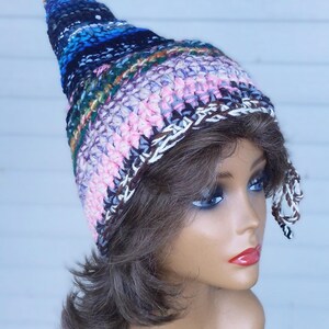 May include: A pointed crochet hat featuring a multi-colored striped pattern in shades of blue, pink, green, and brown. The hat has a tall, conical shape with a textured yarn finish and braided ties hanging from the sides near the bottom edge.