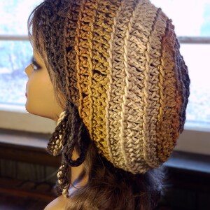 May include: A handmade crochet hat in a gradient of brown, tan, and gold hues. The hat is shown on a mannequin head, with a textured stitch pattern and dangling yarn accents. The hat is a stylish accessory.