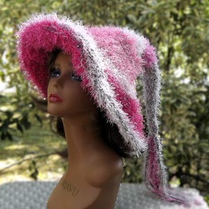 May include: A pink and white fuzzy crocheted hat with a long tail. The hat is on a mannequin head.