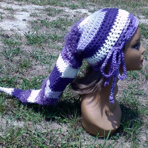 May include: A long, pointed crochet hat featuring horizontal stripes in shades of purple, lavender, and white. The hat has a textured, fuzzy yarn section near the top and includes two braided ties hanging from the sides of the head opening.