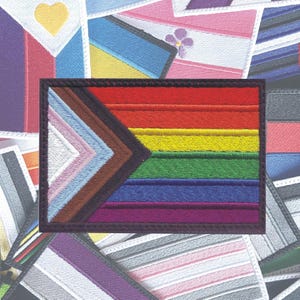 May include: A close-up of a rainbow pride flag with a brown, light blue, pink, and white chevron design in the upper left corner. The flag is embroidered and has a black border.