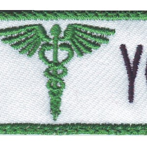 May include: White embroidered name tag with a green border. The tag features a green medical symbol with wings and the text "YOUR NAME HERE".