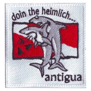 May include: A white embroidered patch with a red and white scuba diving flag. Two cartoon sharks are shown performing the Heimlich maneuver on each other. The text "doin the heimlich..." is above the sharks and the text "antigua" is below the sharks.