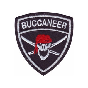 May include: A black and white embroidered patch with the word "BUCCANEER" at the top. The patch features a skull wearing a red bandana, with crossed swords behind it. The patch is shield-shaped.