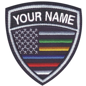 May include: A black and white embroidered patch with a shield shape. The shield features a stylized American flag with thin stripes of red, white, blue, yellow, green, and black. The text "YOUR NAME" is embroidered in white at the top of the shield.