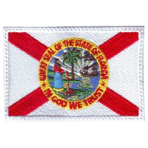 May include: Embroidered patch featuring the Florida state flag. The flag is white with a red diagonal cross and the state seal in the center. The seal depicts a sun rising over a palm tree and the words "Great Seal of the State of Florida" and "In God We Trust".