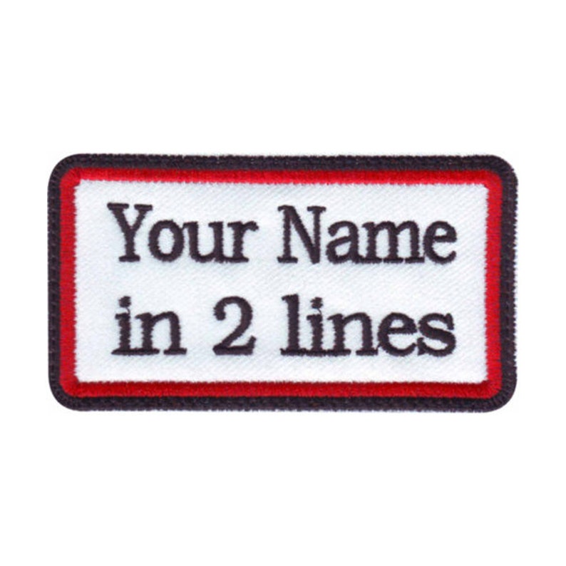 Op de afbeelding: Witte rechthoekige patch met een rode rand en zwarte stiksels. De tekst "Your Name in 2 lines" is in zwart geborduurd.