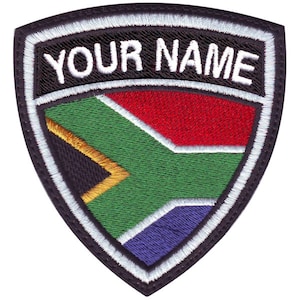 May include: A black and white embroidered patch with a shield shape. The shield features the South African flag in the center and the text "YOUR NAME" at the top.