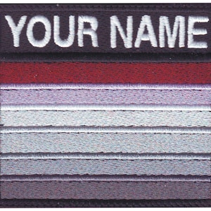May include: A black and white embroidered patch with the text "YOUR NAME" in white. The patch has a red stripe at the top and a gradient of grey stripes below.