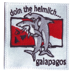 May include: Embroidered patch with a white background and black outline. The patch features a red and white scuba diving flag with two grey sharks performing the Heimlich maneuver. The text "doin the heimlich..." is above the flag and "galapagos" is below the flag.