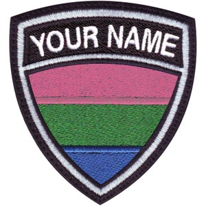 May include: A black and white embroidered patch with a shield shape. The shield has three horizontal stripes in pink, green, and blue. The text "YOUR NAME" is embroidered in white on a black banner above the shield.
