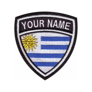May include: A black and white embroidered patch with the flag of Uruguay. The flag features a sun with a face and nine horizontal blue and white stripes. The patch has a black border and the text "YOUR NAME" in white.