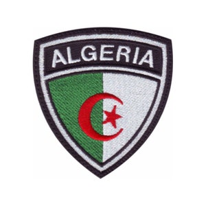May include: A black and white embroidered patch with a shield shape. The shield has a green and white background with a red crescent moon and star. The word "ALGERIA" is embroidered in white on a black background at the top of the shield.
