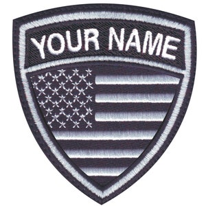 May include: Black and white embroidered patch with a shield shape. The shield features a white American flag with stars and stripes. The top of the shield has a white border with the text "YOUR NAME" in white.