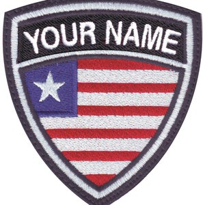 May include: A black and white embroidered patch with a shield shape. The shield has a white star and red and white stripes resembling an American flag. The text "YOUR NAME" is embroidered in white on a black background at the top of the shield.