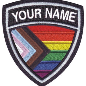 May include: A black and white embroidered patch with a shield shape. The shield features a rainbow flag with additional colors, including brown, light blue, and pink. The top of the shield has a white border with the text "YOUR NAME" in black.