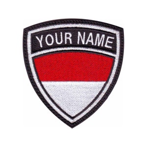 Peut inclure: Un écusson brodé en noir et blanc avec un bouclier rouge, blanc et noir. Le bouclier porte le texte "YOUR NAME" en lettres blanches sur fond noir. Le bouclier est entouré d'une bordure noire.