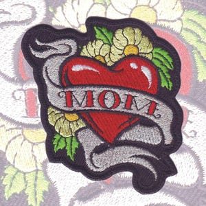 May include: Embroidered iron-on patch with a red heart and a banner that says "MOM". The heart is surrounded by green leaves and white flowers.