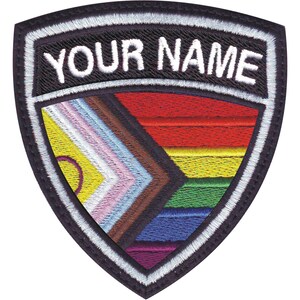 May include: A black and white embroidered patch in the shape of a shield with the text "YOUR NAME" at the top. The shield features a colorful design with stripes of red, orange, yellow, green, blue, purple, brown, and pink.