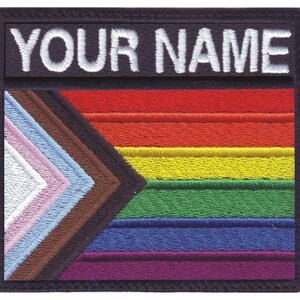 May include: A black and white embroidered patch with the text "YOUR NAME" above a rainbow flag with brown, light blue, pink, white, green, yellow, orange, and red stripes.
