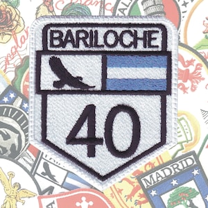 May include: White embroidered patch with a black outline. The patch features a black silhouette of a bird, a blue and white striped flag, and the text "BARILOCHE" and the number "40".