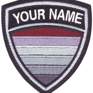 May include: An embroidered patch in the shape of a shield, with a black and white design. The shield features a red, white and grey striped pattern. The text "YOUR NAME" is embroidered in white on a black background at the top of the shield.