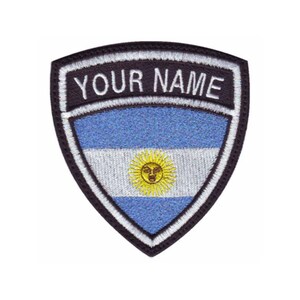 May include: A black and white embroidered patch with a shield shape. The shield features the flag of Argentina with a blue and white striped background and a yellow sun in the center. The text "YOUR NAME" is embroidered in white on a black background above the flag.